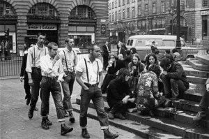 A group of men wearing skinhead suspenders walking by people sitting on the stairs in the city during the 1960's
